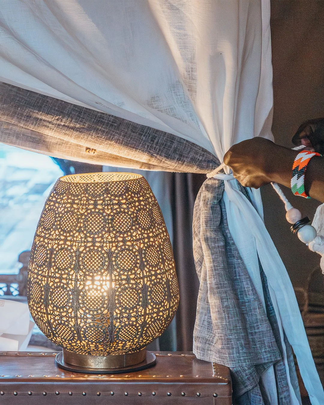 A hand drawing back a curtain beside a patterned brass lamp
